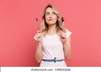 Portrait of thoughtful blond woman 20s wearing body measuring tape around waist holding spoon and fork isolated over pink background