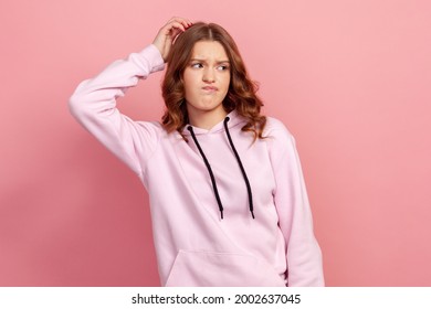 Portrait Of Thoughtful Confused Brunette Girl In Hoodie Scratching Head Having Doubts, Not Sure Contemplating With Uncertain Puzzled Face. Indoor Studio Shot Isolated On Pink Background