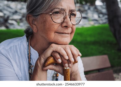 Portrait of a thoughtful elderly woman of 70 years old sitting on a bench in the park leaning on a stick due to walking disability, the elderly lady looks away - Powered by Shutterstock