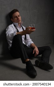 Portrait of a thoughtful handsome young man with a glass of alcohol in his hands sitting on the floor against a stone wall.