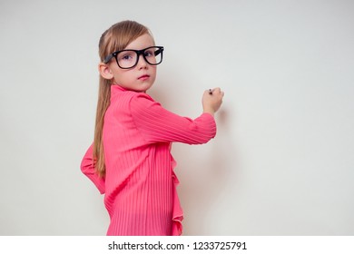 Portrait of thoughtful little smiling girl in a pink blouse and glasses writing on wall blackboard pencil up copyspace. genius child with the idea on white background in studio copy space