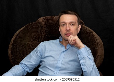 Portrait Of A Thoughtful Man 30-35 Years Old In A Blue Shirt, Sitting In An Armchair On A Black Background. Perhaps He Is A Lawyer Or A Lawyer, A Coach Or A Psychologist.