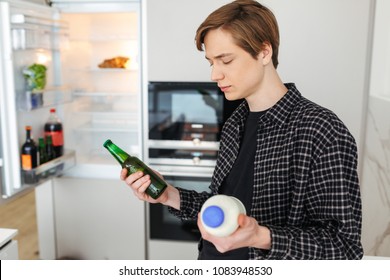 Portrait Of Thoughtful Man Standing Near Fridge With Beer And Milk In Hands At Home Isolated