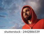 Portrait of a thoughtful man in a vibrant orange hoodie, gazing into the distance under a dramatic sky.