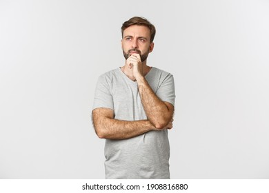 Portrait of thoughtful modern guy in grey t-shirt, making choice, looking at upper left corner and thinking, standing over white background