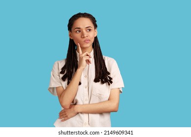 Portrait Of Thoughtful Woman With Black Dreadlocks Thinking About Future, Holding Chin, Having Serious Facial Expression, Wearing White Shirt. Indoor Studio Shot Isolated On Blue Background.