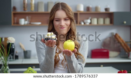 Portrait of thoughtful woman choosing between healthy food and unhealthy food in kitchen. Closeup of smiling girl looking at dessert and apple indoors. Emotional woman preferring fresh fruit