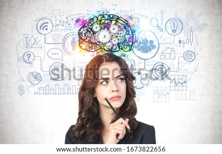 Portrait of thoughtful young businesswoman with pencil and long wavy hair standing near concrete wall with colorful brain sketch and business icons drawn on it. Concept of planning