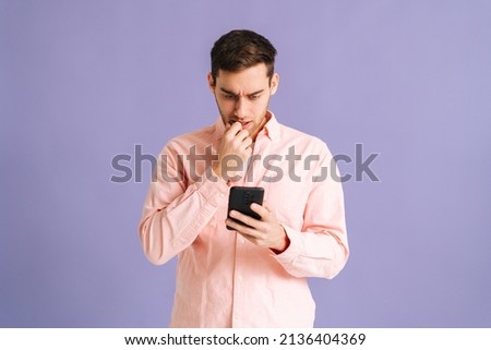 Portrait of thoughtful young man holding phone with doubtful and skeptical expression on pink isolated background. Studio shot of handsome guy thinking and reading online message using smartphone.