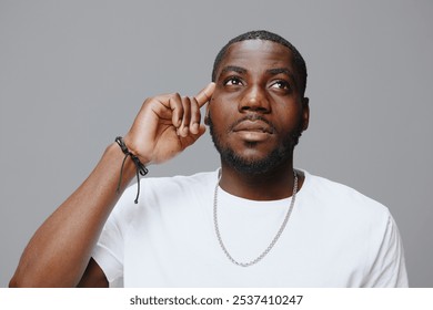 Portrait of a thoughtful young man wearing a white t shirt, looking upwards with a hand on his temple, expressing curiosity and contemplation against a neutral gray background - Powered by Shutterstock