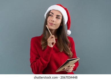 Portrait of thoughtful young woman in Santa's hat holding copybook and pencil in hands ready to write Christmas wish list or holiday greetings.