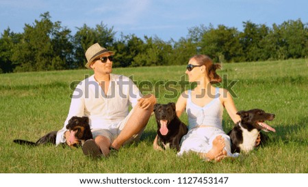 PORTRAIT: Three adorable black puppies rest by their cheerful owners' sides in the middle of a sunny meadow. Smiling young girlfriend and boyfriend hold and rub their lovely young border collie dogs.