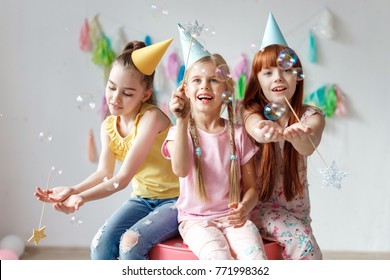 Portrait of three beautiful girls wear festive caps, play with bubbles, sit together on chair, celebrate birthday, being in good mood, use magic wand, have party in decorated room. Childhood concept
