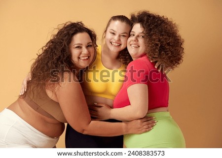 Portrait of three beautiful young women with overweight body standing in sportswear, smiling over beige studio background. Concept of sport, body-positivity, weight loss, body and health care