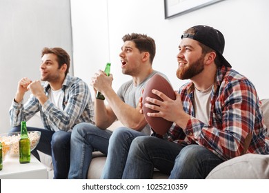 Portrait Of Three Cheerful Young Men Watching American Football While Sitting At Home With Beer And Snacks Indoors