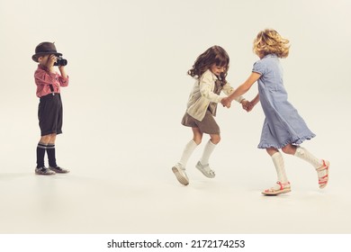 Portrait Of Three Children, Little Boy Taking Photo Of Two Cheerful Girls Holding Hands And Whirling Isolated Over Grey Background. Concept Of Childhood, Friendship, Family, Fun, Lifestyle, Fashion