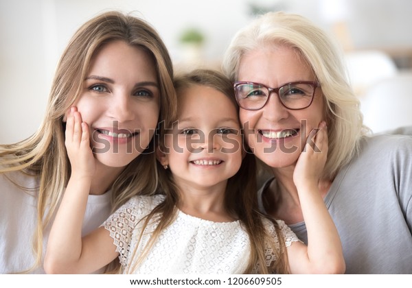 Portrait of three generations of women look at\
camera posing for family picture, cute little girl hug mom and\
granny enjoy time at home, smiling mother, daughter and grandmother\
spend weekend together