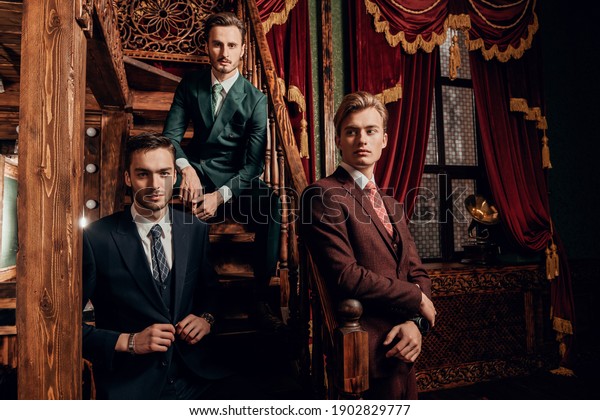 Portrait of three handsome men in elegant\
classic suits posing in a luxury apartments with classic interior.\
Men\'s beauty, fashion.