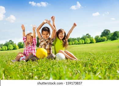 Portrait of three happy kids, boy and girls sitting in the grass in park with lifted hands and holding sport balls