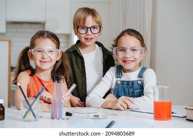 Portrait Of Three Happy Little Kids Doing Home Science Project, All Behid Table. All Wearing Glasses. Chemical Glassware And Colored Liquids On The Table.