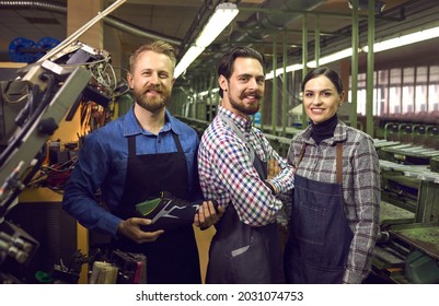 Portrait of three happy proud satisfied young shoe factory workers standing together in assembly line workshop, smiling and looking at camera. Footwear manufacturing industry concept
