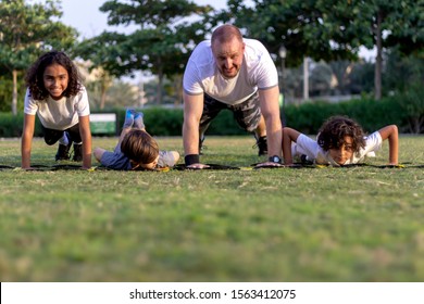 Portrait Of Three Happy Smiling Kids And Their Coach Doing Push-up Exercises Standing In A Row Outdoor On The Grass.Mixed Group Of People Attending Fitness Class,doing Push Ups On The Green Field.