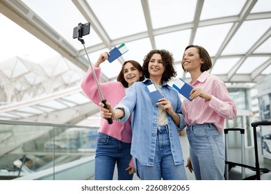 Portrait Of Three Happy Women Taking Photo Together At Airport, Cheerful Young Female Friends Using Smartphone And Selfie Stick, Smiling Traveller Ladies Holding Passports And Posing At Camera - Powered by Shutterstock