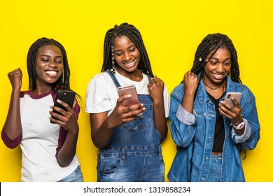 Portrait Of Three Happy Young African Woman Friends Smiling While Using Mobile Phone Together With Win Gesture On Yellow Background