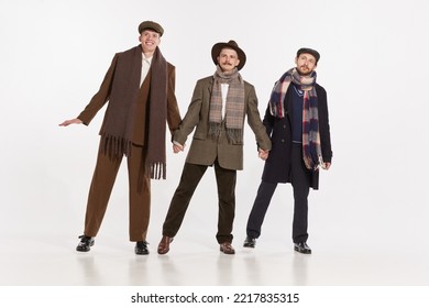 Portrait Of Three Stylish Men In Warm Coats, Scarves And Hats Posing Over Grey Studio Background. Male Fashion. Concept Of Retro Fashion, Style, Friendship, Creativity, Youth Culture, Emotions, Ad