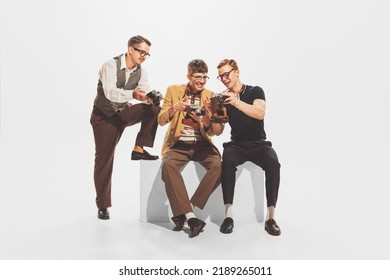 Portrait Of Three Stylish Young Men In Trendy Suits Sitting, Looking At Retro Camera Photos Isolated Over White Studio Background. Concept Of Retro Fashion, Style, Youth Culture, Emotions, Art, Ad