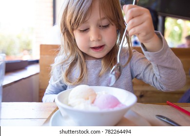 Portrait Of Three Years Old Blonde Child Ready To Eat Two Scoops Of Strawberry And Vanilla Ice Cream, In White Bowl, With A Spoon In Raised Hand, Sitting At The Restaurant Table