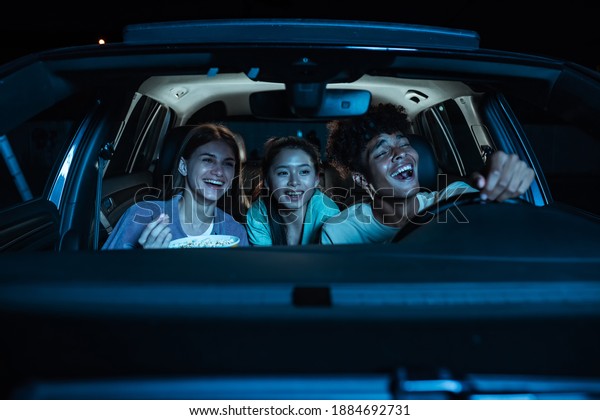 Portrait of three young friends looking emotional,
laughing while sitting together in the car and watching a movie in
a drive in cinema. Entertainment, leisure activities concept.
Horizontal shot