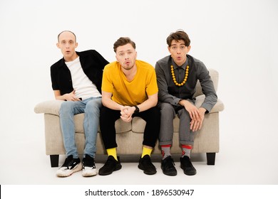 Portrait Of Three Young Friends Spend Time Together Sitting On The Small Sofa Looking At The Camera Isolated On White Background. Group Of Guys Sitting On Couch And Communicates On White.