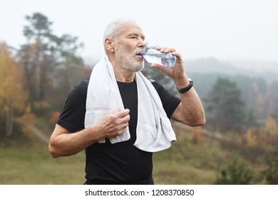 Portrait Of Tired Active Mature Sixty Year Old Man In Black T-shirt Drinking Water From Plastic Bottle While Running Outdoors, Foggy Autumn Forest In Background. Sports, Activity And Aging Concept