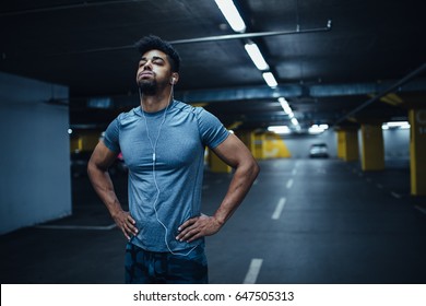 Portrait of a tired african american athlete man holding his hands on hips and listening to music in the underground car parking. - Powered by Shutterstock