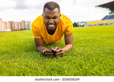 Portrait Of A Tired African American Male Working Out Hard On A Grass Field At The Stadium. Young Athletic Man Doing Plank Exercise Outdoors In Yellow Sportswear, Training Concept