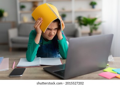 Portrait Of Tired Angry Student Girl Putting And Holding Notebook On Her Head Looking Angrily At Laptop Screen, Teen Studying Remotely At Home. Stressed Mad Furious Pupil Tired Of Homework