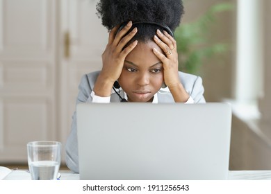 Portrait of tired annoyed afro businesswoman, wear headphones, talking to a bad customer looking at screen laptop in office. Office employee confused by message of a disgruntled buyer, touching head