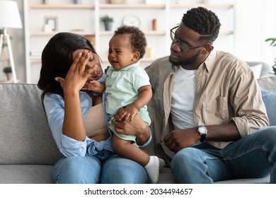 Portrait Of Tired Black Mum Sitting With Her Small Crying Black Kid On Couch At Home, Feeling Headache And Migraine, Touching Forehead With Hand, Sad Dad Comforting Her. Exhaustion And Parenthood