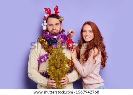 Portrait of tired boyfriend decorated with Christmas baubles and tinsel, happy woman playing with angry man, teasing and kissing, expressing happiness