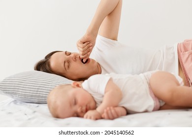 Portrait of tired exhausted mother lying on bed and yawning, covering open mouth with fist, wakes up early while her baby girl sleeping, posing at home in light room.