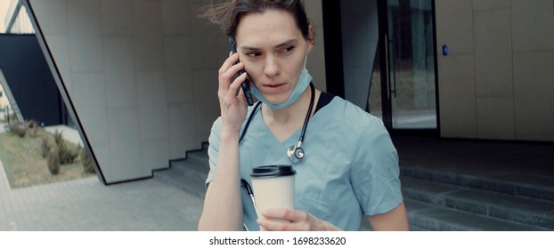 Portrait Of Tired Exhausted Nurse Or Doctor Having A Phone Call Outside In The Morning. COVID-19, Coronavirus Pandemic