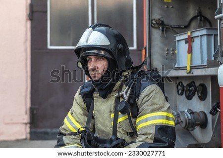 Portrait of a tired firefighter in a protective suit and a protective helmet sitting by a fire engine after returning to the fire department