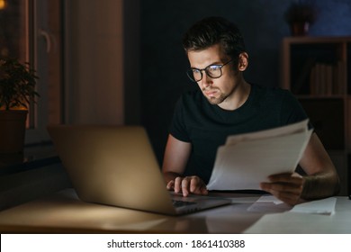 Portrait of tired focused young man in glasses working remotely late at night at home office with computer, looking at laptop, thinking about deadline. Overwork, negative thoughts, deadline concept - Shutterstock ID 1861410388