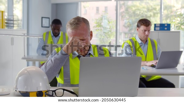 Portrait of tired industrial\
architect working on laptop in office. Frustrated and stressed\
engineer sitting at desk in workplace having problem with\
project