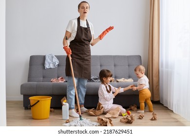 Portrait Of Tired Mother Wearing Brown Apron Cleaning At Home, Woman With Mop Cleaning With Kids Sitting On Floor At The Sofa, Exhausted Housewife Of Tidying Up House.