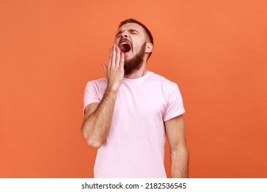 Portrait of tired sleepless bearded man yawning and covering mouth with hand, feeling exhausted, lack of sleep, wearing pink T-shirt. Indoor studio shot isolated on orange background.