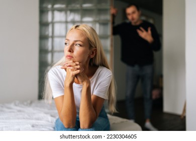 Portrait of tired stressed woman sadness looking away sitting on bed on blurred background of aggressive husband shouting on wife at home. Concept of family scandal, crisis, domestic violence, abuse. - Shutterstock ID 2230304605
