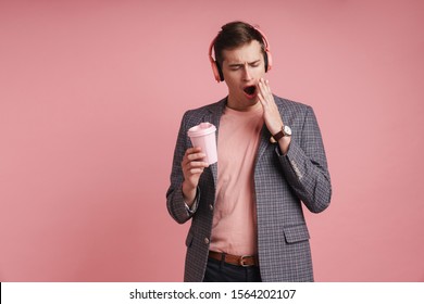 Portrait of a tired young attractive man wearing jacket standing isolated over pink background, lstening to music with wireless headphones, holding coffee cup