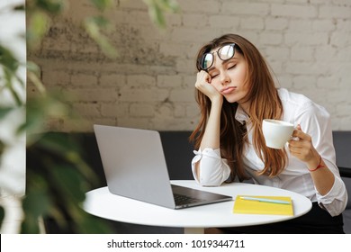Portrait of a tired young businesswoman sitting at the table with laptop computer while holding cup of coffee and sleeping at a cafe